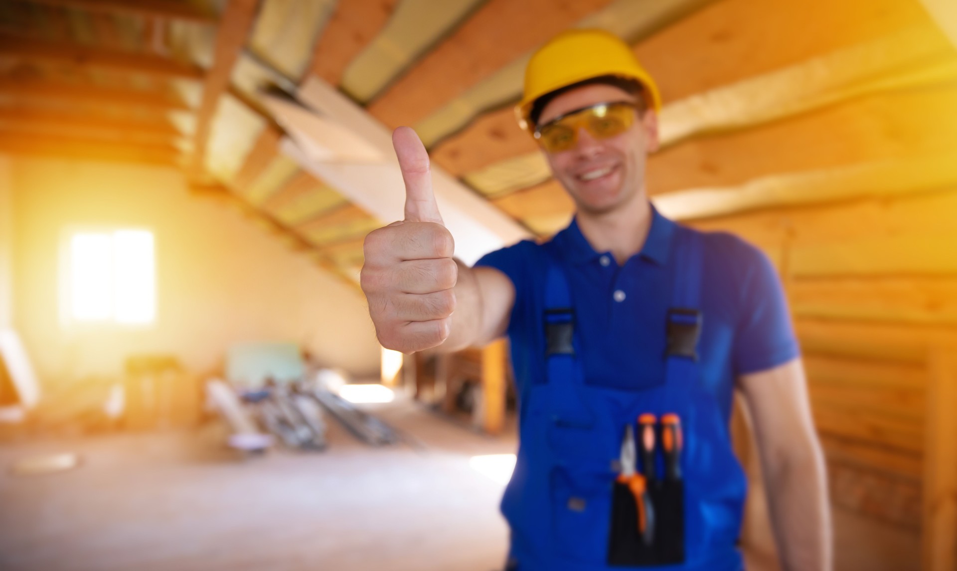 Good work. Builder in a yellow hard hat and safety glasses gives a thumbs-up in an unfinished attic. The bright, warm lighting and his smile convey a confident attitude towards the renovation project